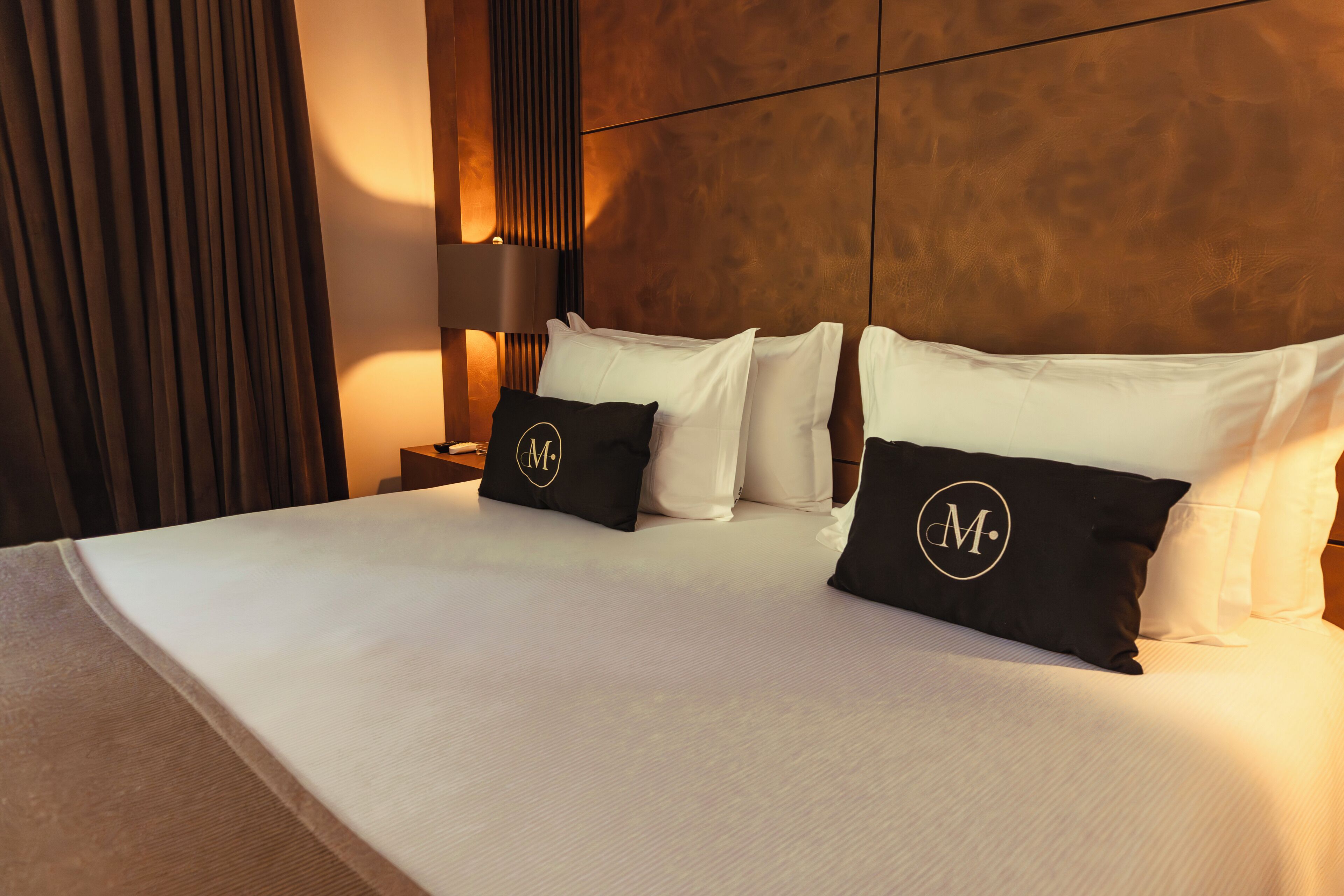 A neatly made bed with white linens and black pillows with a monogram, a bedside table with a lamp, and a wooden headboard.