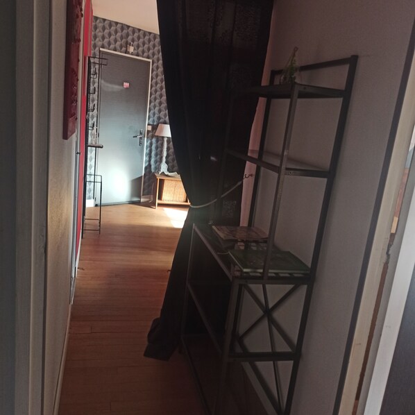 Een smalle gang met een houten vloer, een metalen schap en een gedeeltelijk open deur die leidt naar een kamer met behang met een patroon.