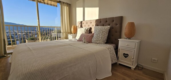 Une chambre avec un grand lit, une table de chevet, une lampe décorative et une vue sur le paysage urbain, visible depuis un balcon.