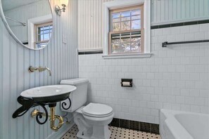 Une salle de bain avec un lavabo, des toilettes et une baignoire, tous de couleur blanche. Il y a une fenêtre au-dessus des toilettes et un miroir au-dessus du lavabo.