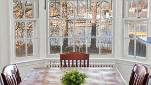 A dining room with a wooden table and chairs, large windows, and a view of trees and a building outside.