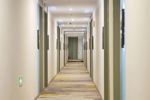 Ein langer, schmaler Hotelkorridor mit Türen an beiden Seiten, einem gemusterten Teppich und einem grünen Notausgangsschild.