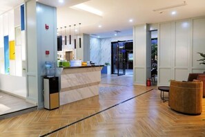 Een hotellobby met een receptiebalie, een waterdispenser, een kleine koelkast, een zithoek met een bank en uitzicht op de eetruimte.