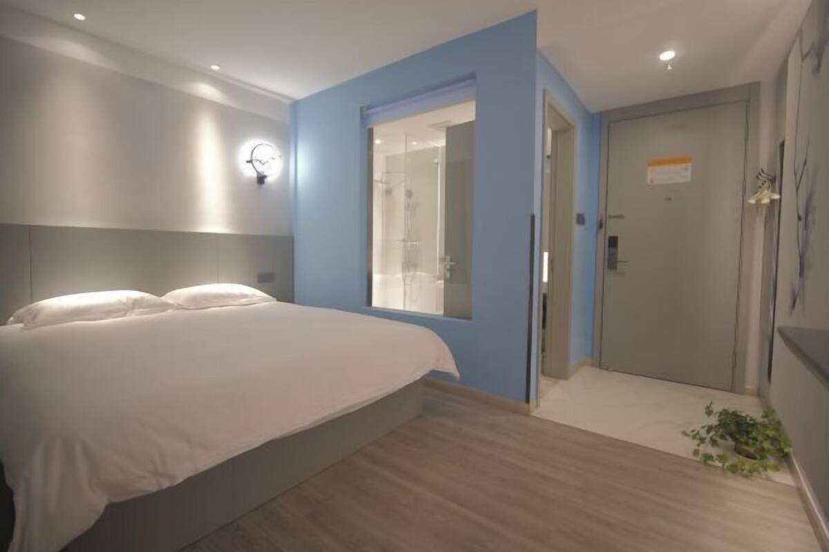 Une chambre à coucher avec un lit, une salle de bain avec une porte en verre et une petite plante posée sur le sol.