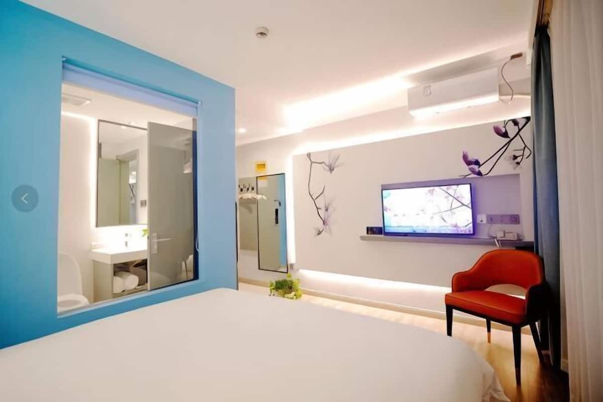 Une chambre à coucher moderne avec un lit, un fauteuil rouge, un téléviseur et une salle de bain visible à travers une porte en verre.