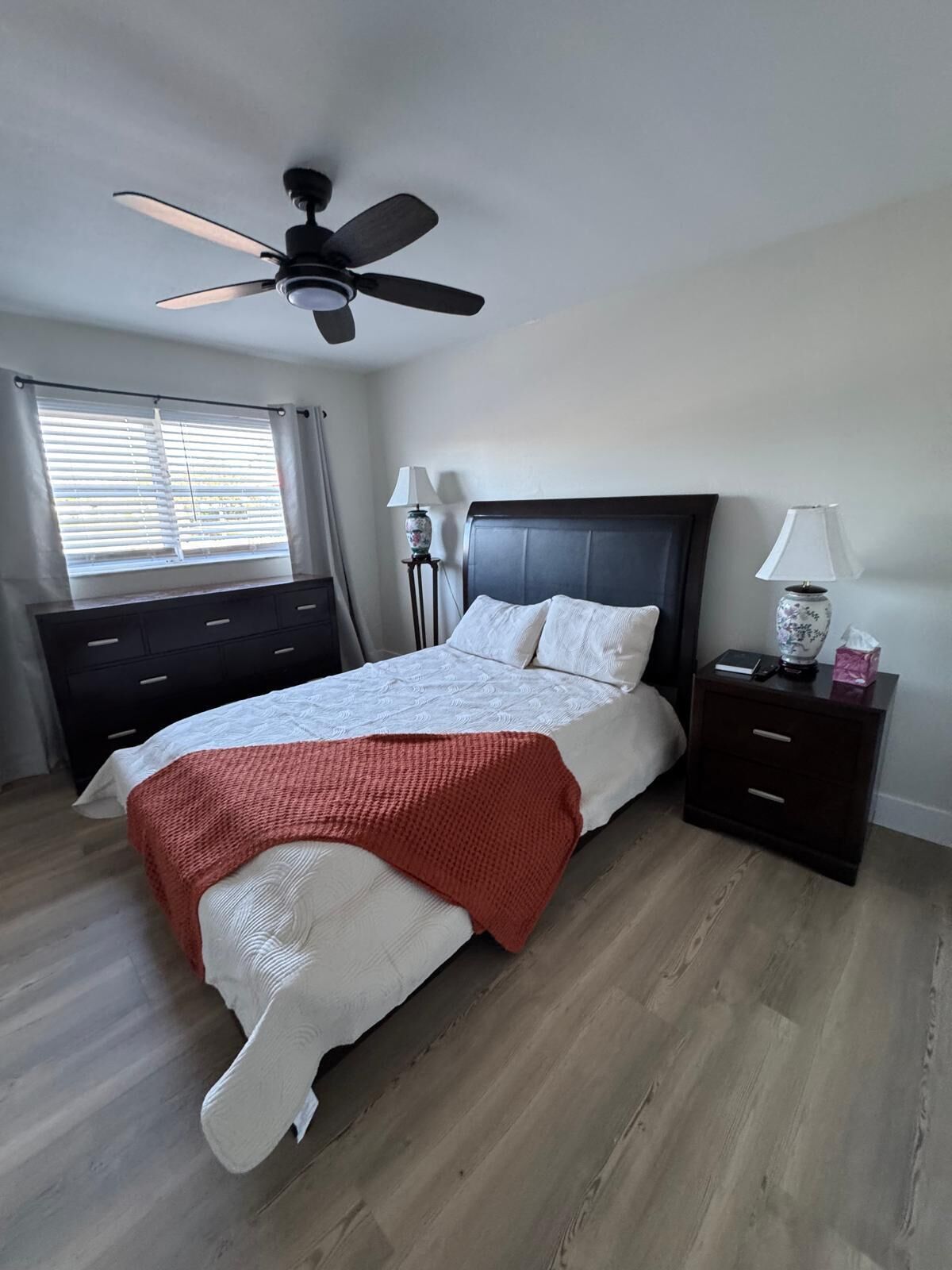 A bedroom with a bed, a ceiling fan, a nightstand, a lamp, and a window with blinds.