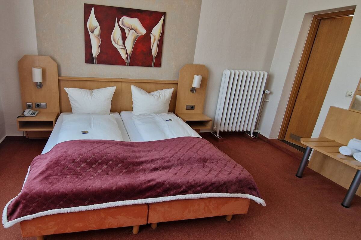 Ein Hotelzimmer mit einem Doppelbett, zwei Nachttischen, einem Heizkörper, einem Spiegel und einem Fenster mit Vorhängen.