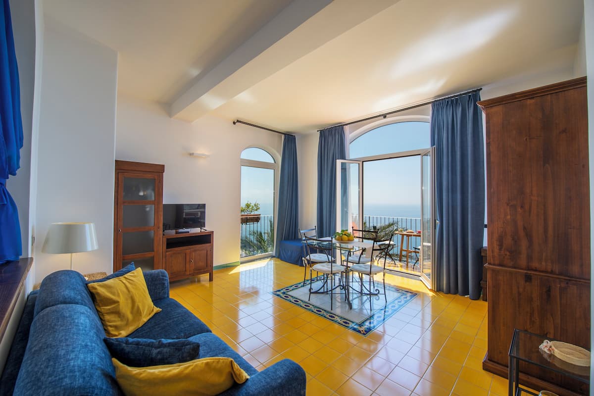 A living room with a sofa, a TV, a wooden cabinet, a dining table with chairs, and a balcony with a view.