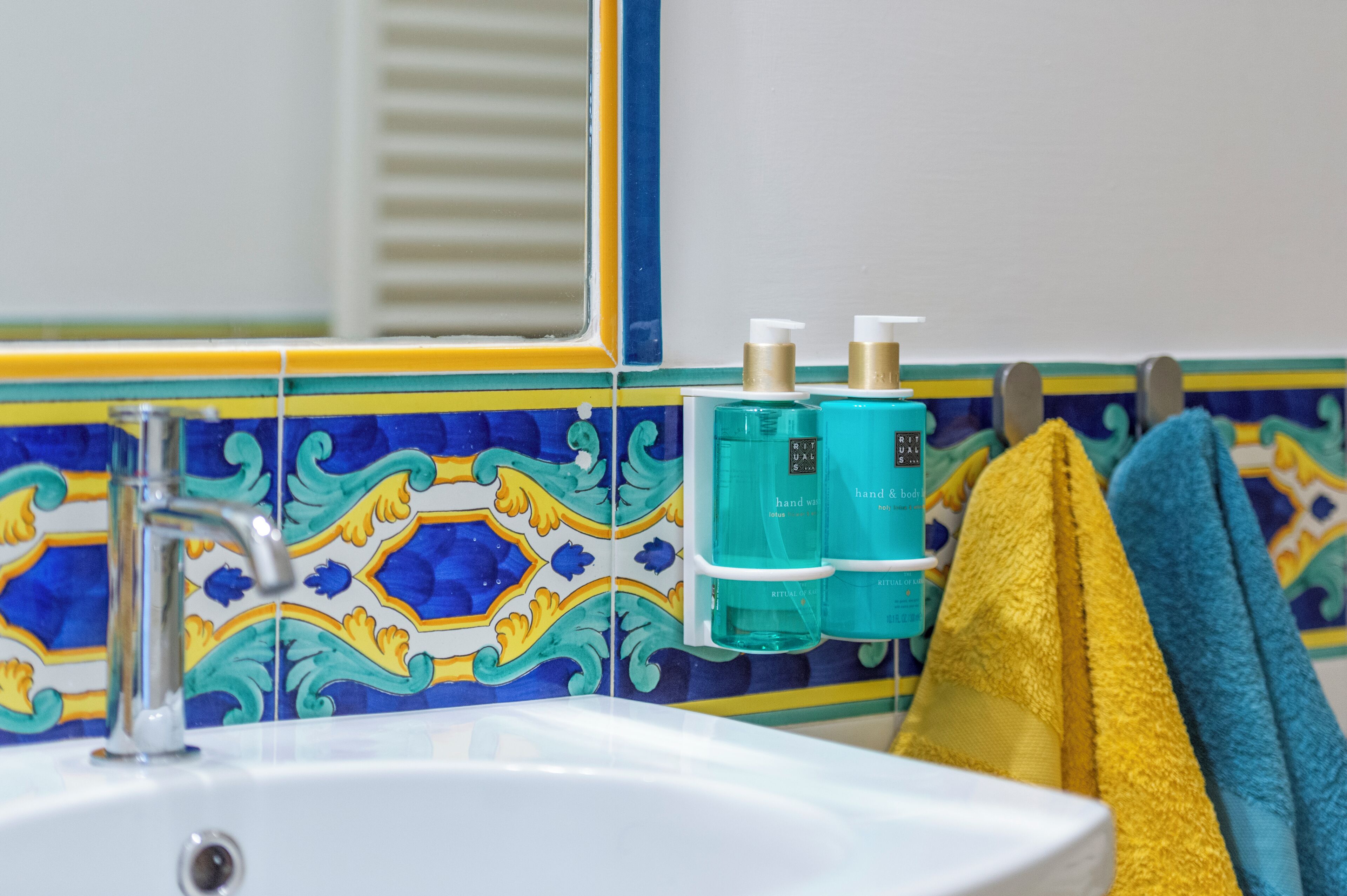 A bathroom with a white sink, a mirror, and a tiled backsplash. There are two bottles of hand soap and a yellow towel hanging on a rack.