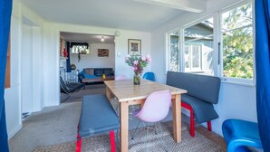 A cozy living area with a wooden table, benches, and a sofa. There is a window with blue curtains, a painting on the wall, and a vase with flowers on the table.