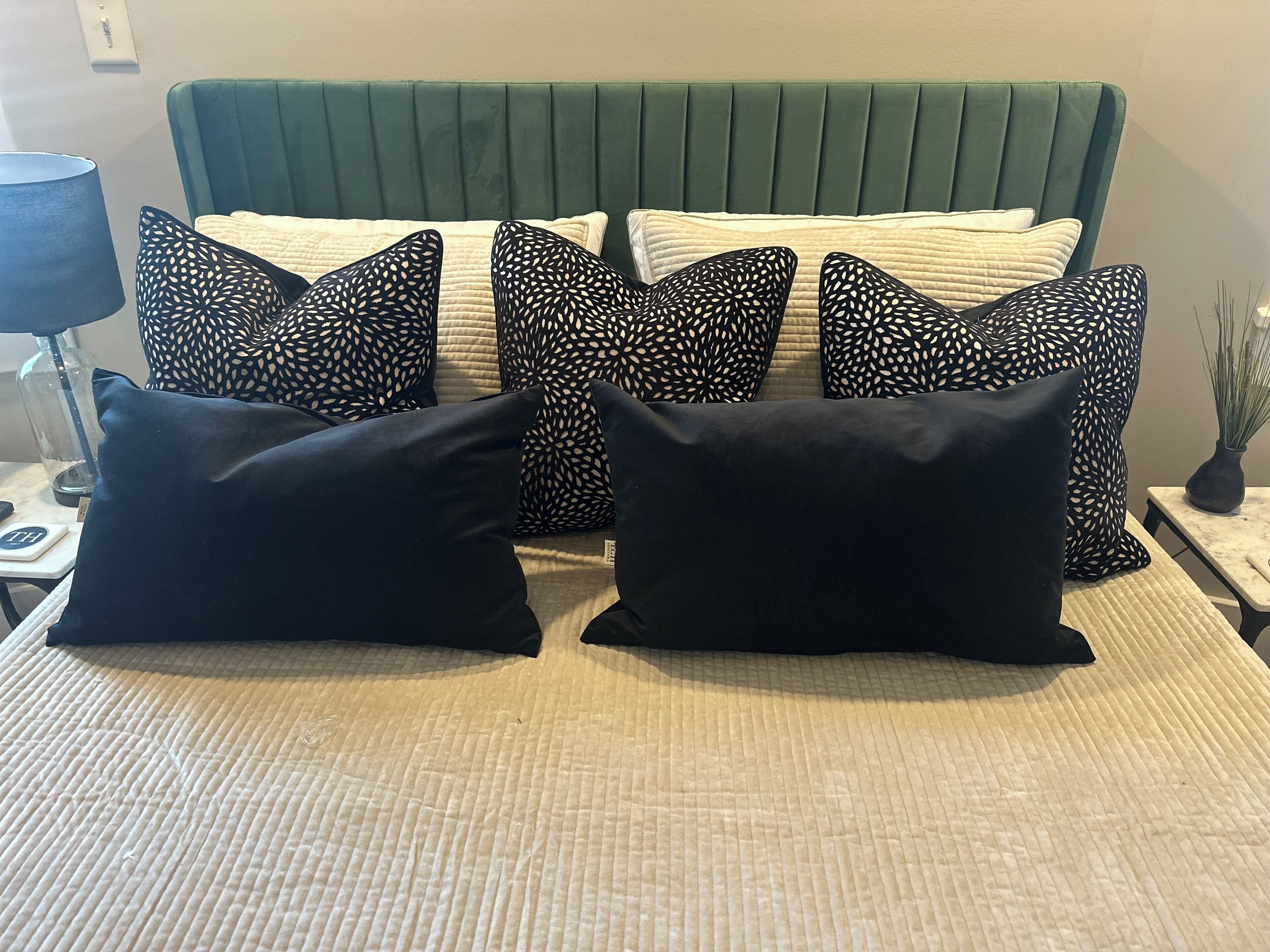 A bed with a green upholstered headboard, multiple patterned pillows, and a bedside table with a lamp and a vase.