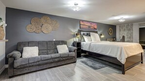 A modern bedroom with a grey sofa, a bed with a white bedspread, a wall-mounted circular decoration, and a painting of a bridge. - Spacious Pool House – 5 Minutes to Elgin Centre (St. Thomas)