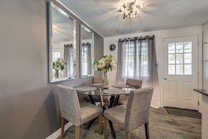 Un coin repas comprenant une table ronde en verre, quatre chaises grises à revêtement rembourré, un vase avec des fleurs et un grand miroir au mur.