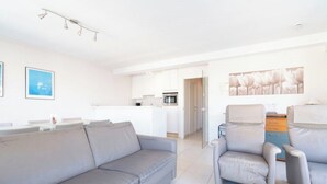A modern living room with a grey sofa, a small table, and a chair. There is a kitchen area in the background with white cabinets and a refrigerator. - Apartment in Middelkerke With Sea View (Middelkerke)