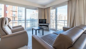 A modern hotel room with a large window, a flat-screen TV, a sofa, and a small table. - Apartment in Middelkerke With Sea View (Middelkerke)