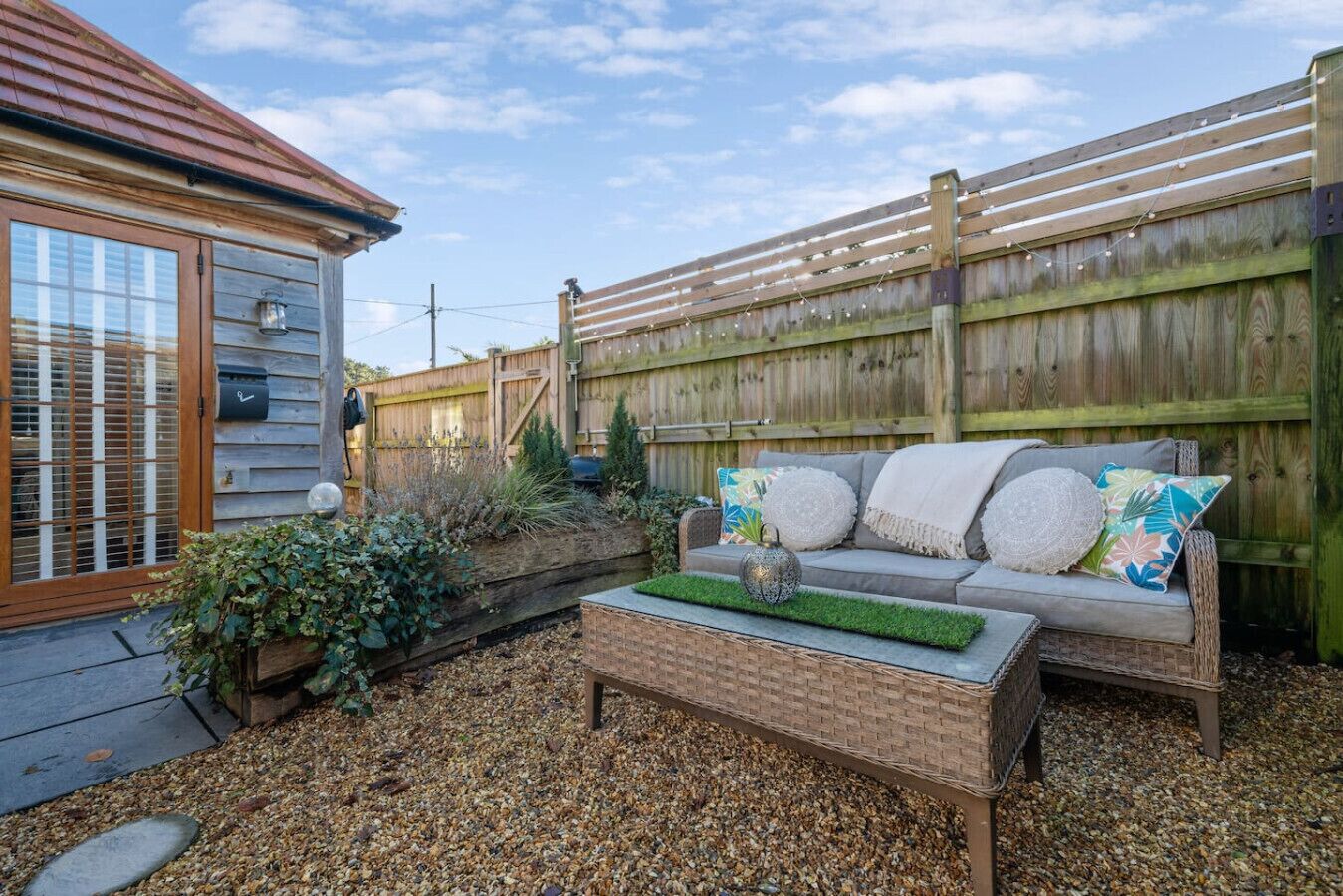 Un jardin à l’arrière de la maison, avec une clôture en bois, un banc en briques, un canapé en osier agrémenté de coussins et un petit espace aménagé en jardin avec des plantes.