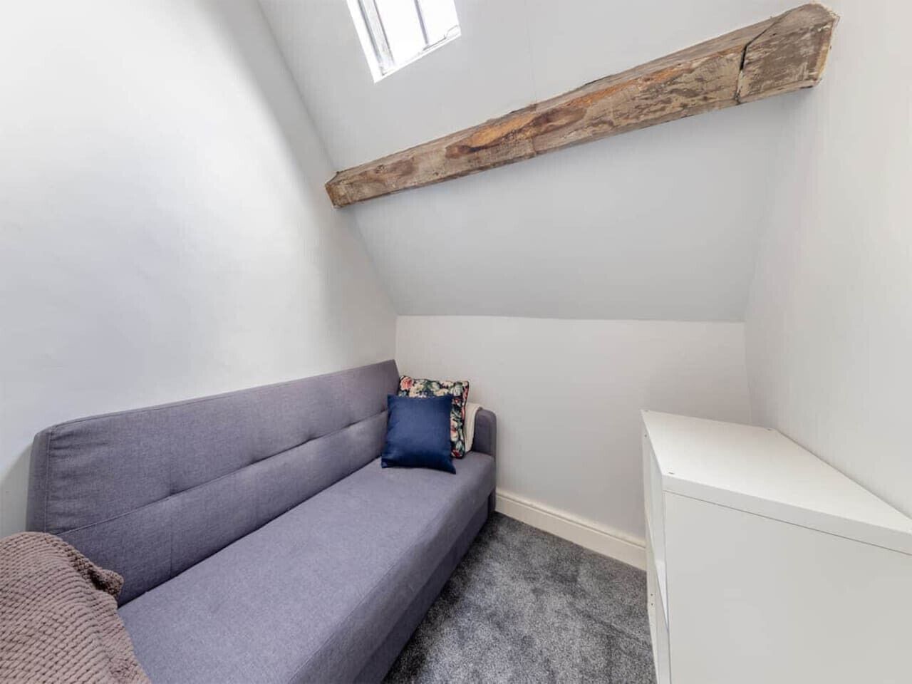 A small, dimly lit room with a grey sofa, a white cabinet, and a wooden beam on the ceiling.