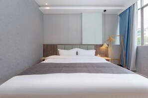 Una habitación de hotel moderna con una cama grande, un cabecero de madera, una mesita de noche con lámpara y una ventana con cortinas.
