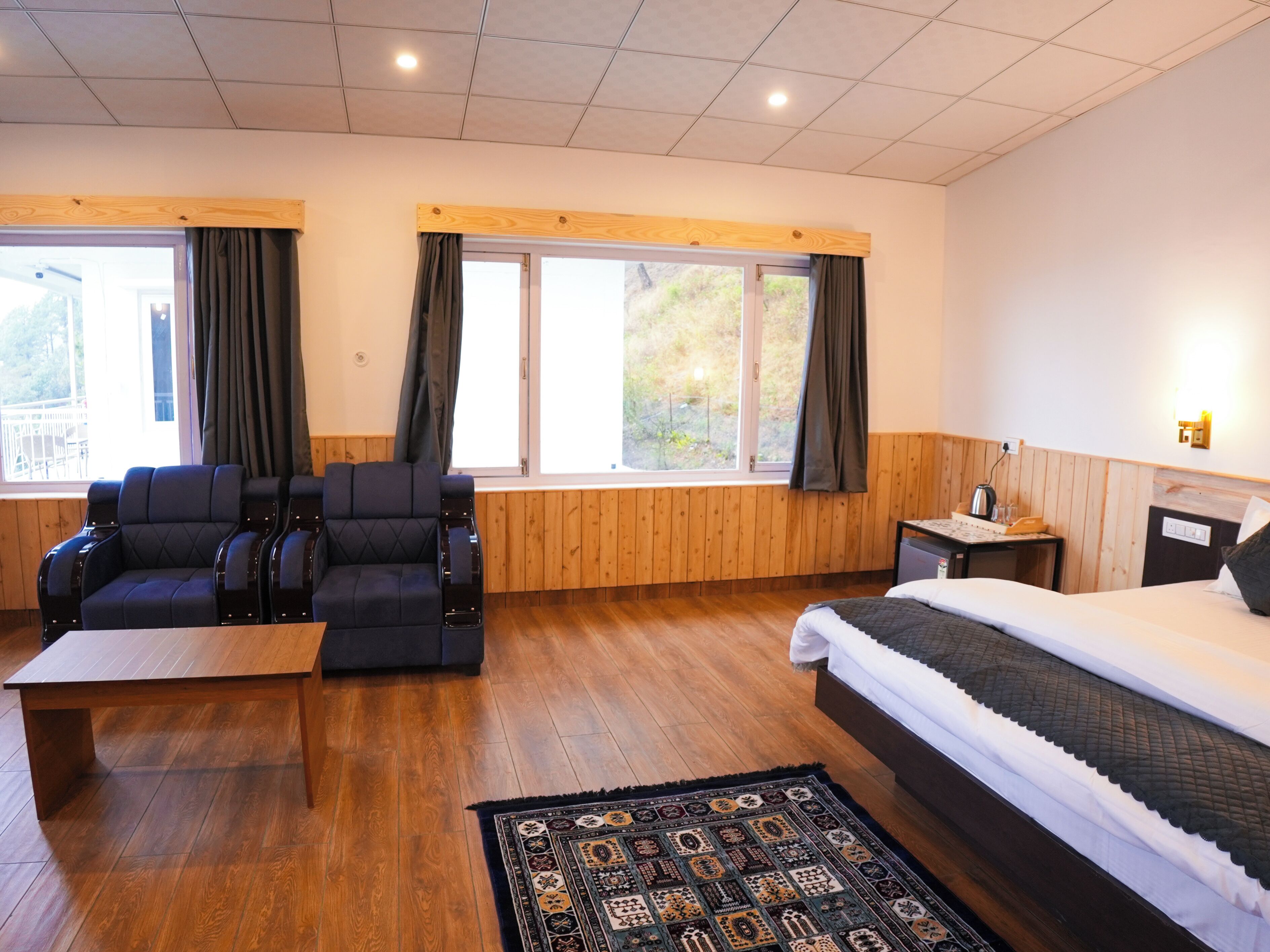 A hotel room with a bed, a sofa, a small table, a nightstand, and two windows with curtains.