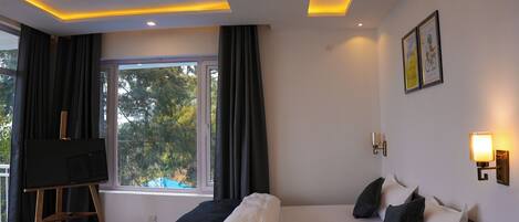 A modern hotel room with a large bed, a desk, a standing fan, and a window with curtains.