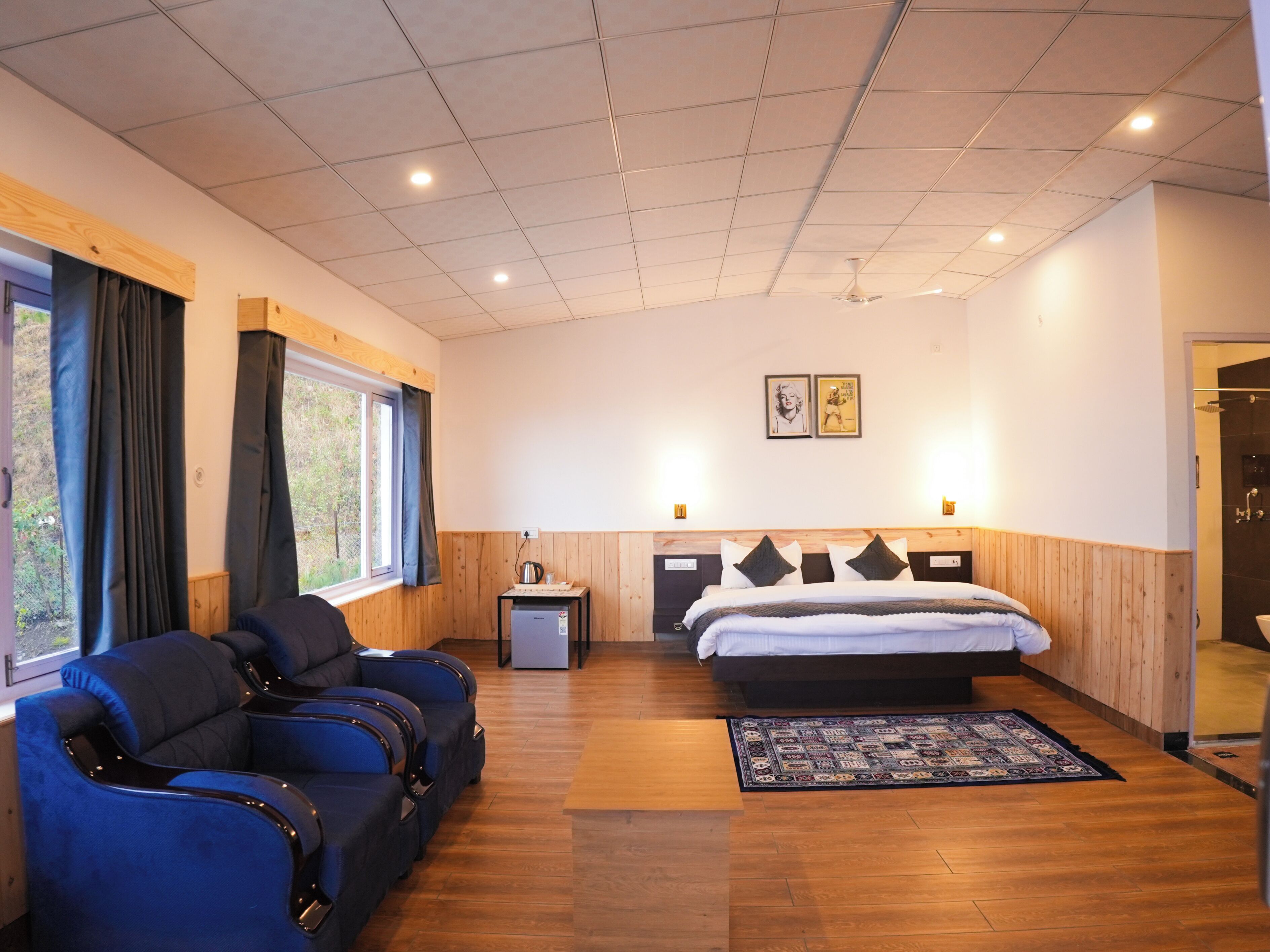Una habitación de hotel moderna con una cama grande, un área de descanso con sillas azules, una mesita con un hervidor y una ventana con cortinas.