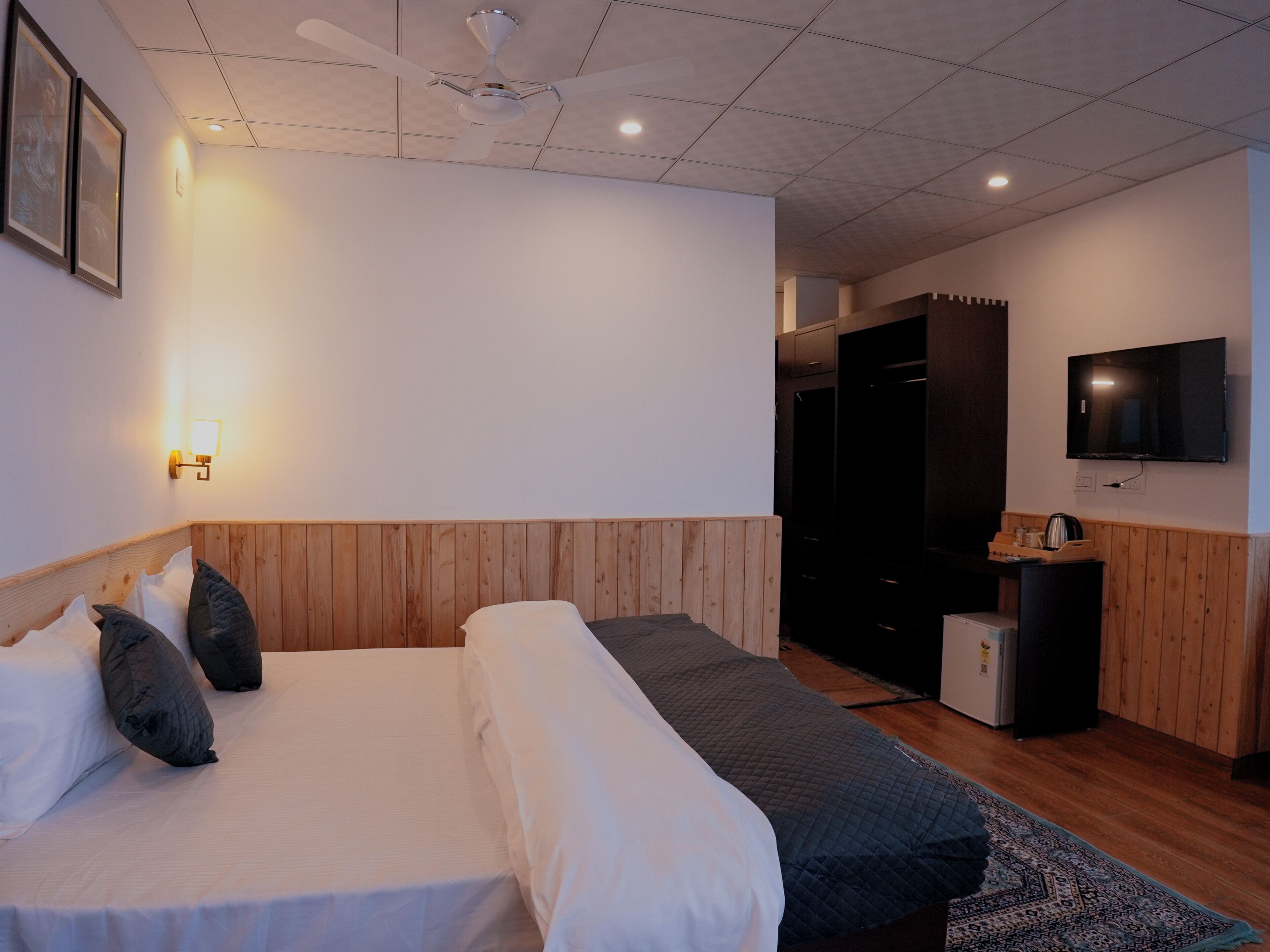 A hotel room with a bed, wooden headboard, a flat-screen TV, a wardrobe, and a small kitchenette.