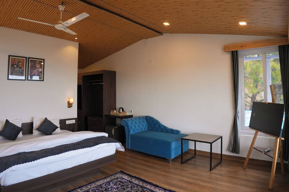 Moderni hotellihuone, jossa on sänky, sininen sohva, pieni pöytä ja maalausteline ikkunan vieressä.