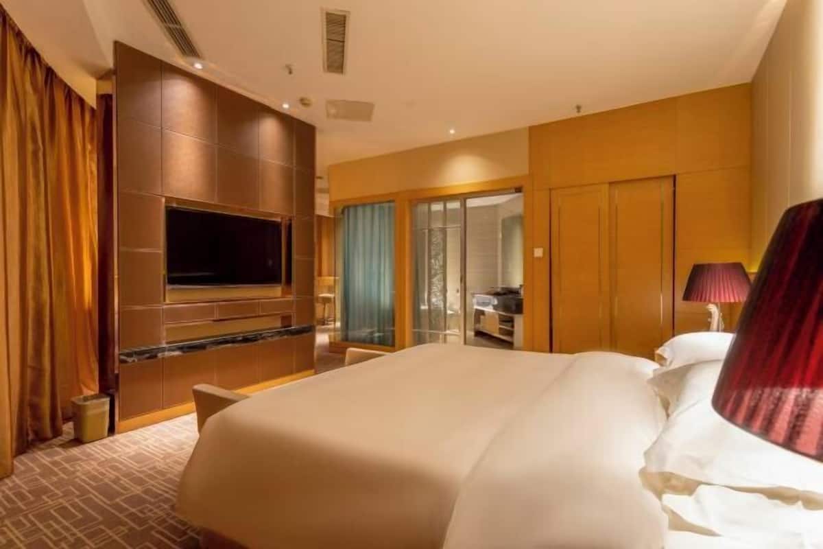 A hotel room with a large bed, a flat-screen TV mounted on the wall, a seating area with a table and chairs, and a sliding glass door leading to a balcony.