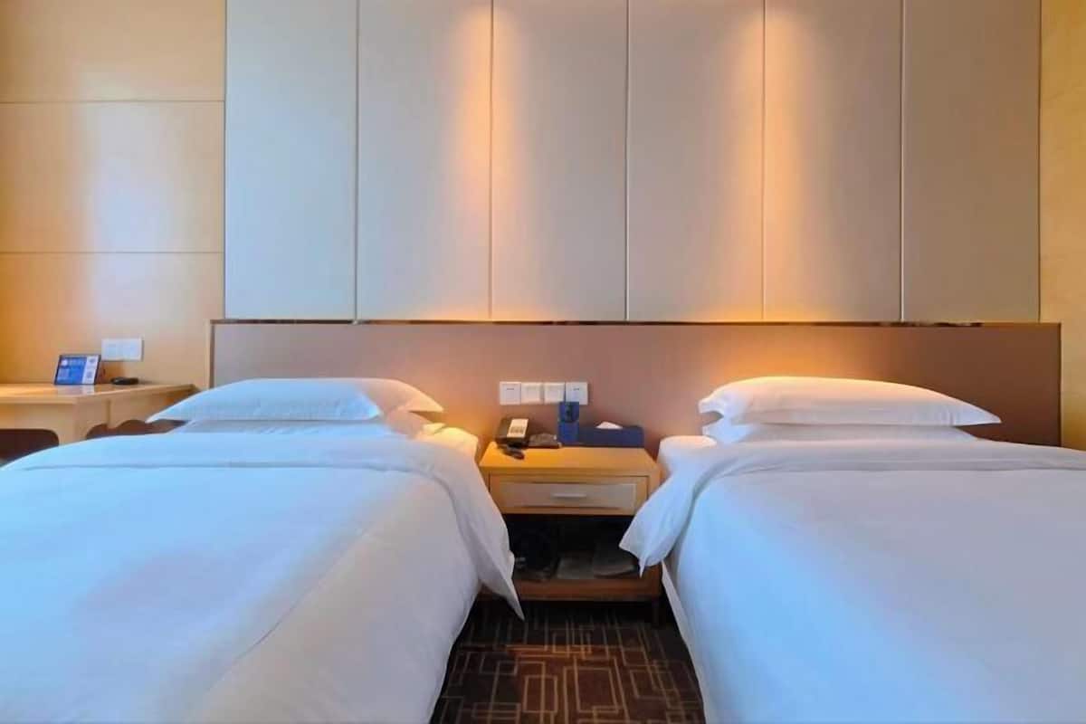 A hotel room with two beds, a nightstand with a phone, and a wall-mounted panel.