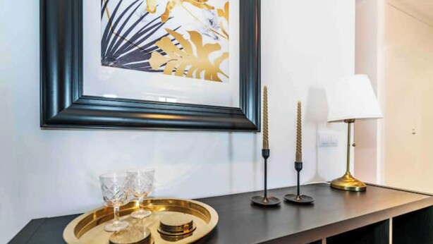 A framed artwork on a wall, a tray with glasses and coins, and a lamp on a dark console table.