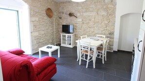 A room with a stone wall, a television on a cabinet, a dining table with chairs, and a red sofa. - Reusia Holiday Home (Ragusa)