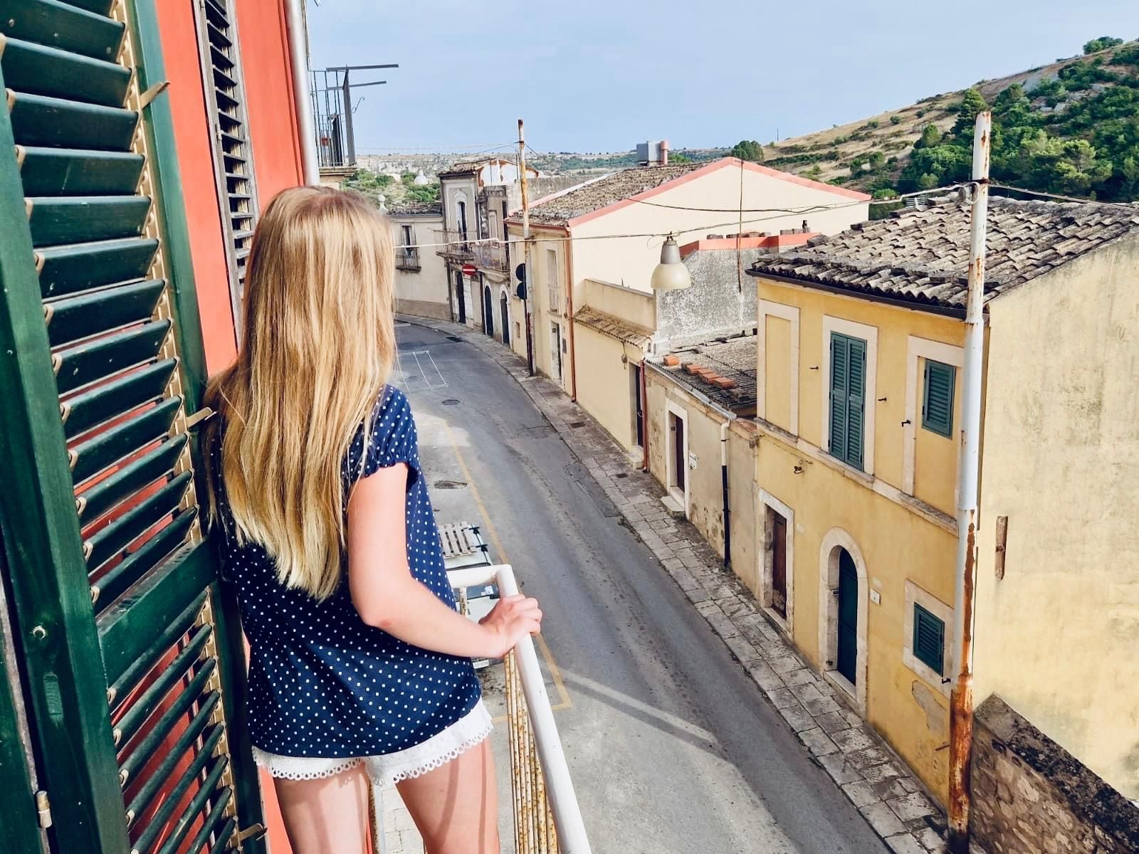 A narrow street with buildings on either side, a clear sky, and a person standing on a balcony.