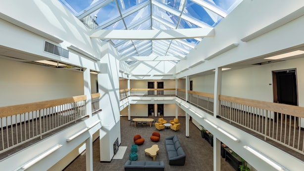 A spacious atrium with a high ceiling and skylight, featuring a central seating area with sofas and tables, surrounded by white walls and wooden railings. - Brewster Hall (Cheney)