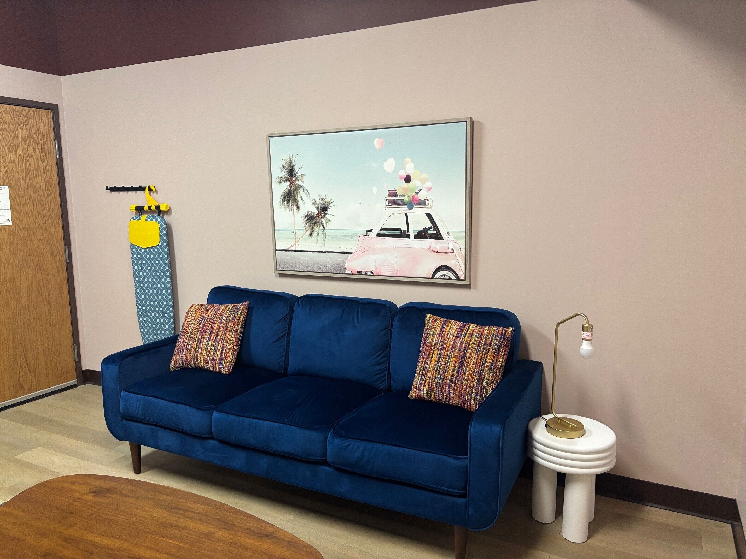 Una habitación con un sofá azul, un cuadro enmarcado de un coche en la playa, un inodoro blanco con asiento dorado y un suelo de madera.
