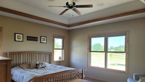 Un dormitorio con una cama grande, un banco de madera, un ventilador de techo y dos ventanas con vistas al exterior.