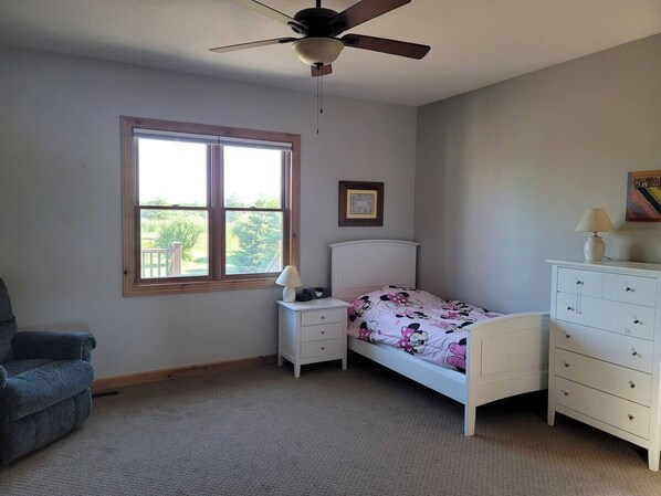 A bedroom with a single bed, a nightstand, a dresser, a chair, a ceiling fan, and a window with a view of greenery.