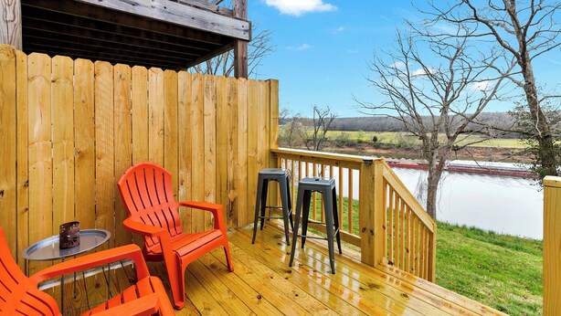 Eine Holzterrasse mit zwei orangefarbenen Stühlen und zwei schwarzen Hocker, mit Blick auf einen See.