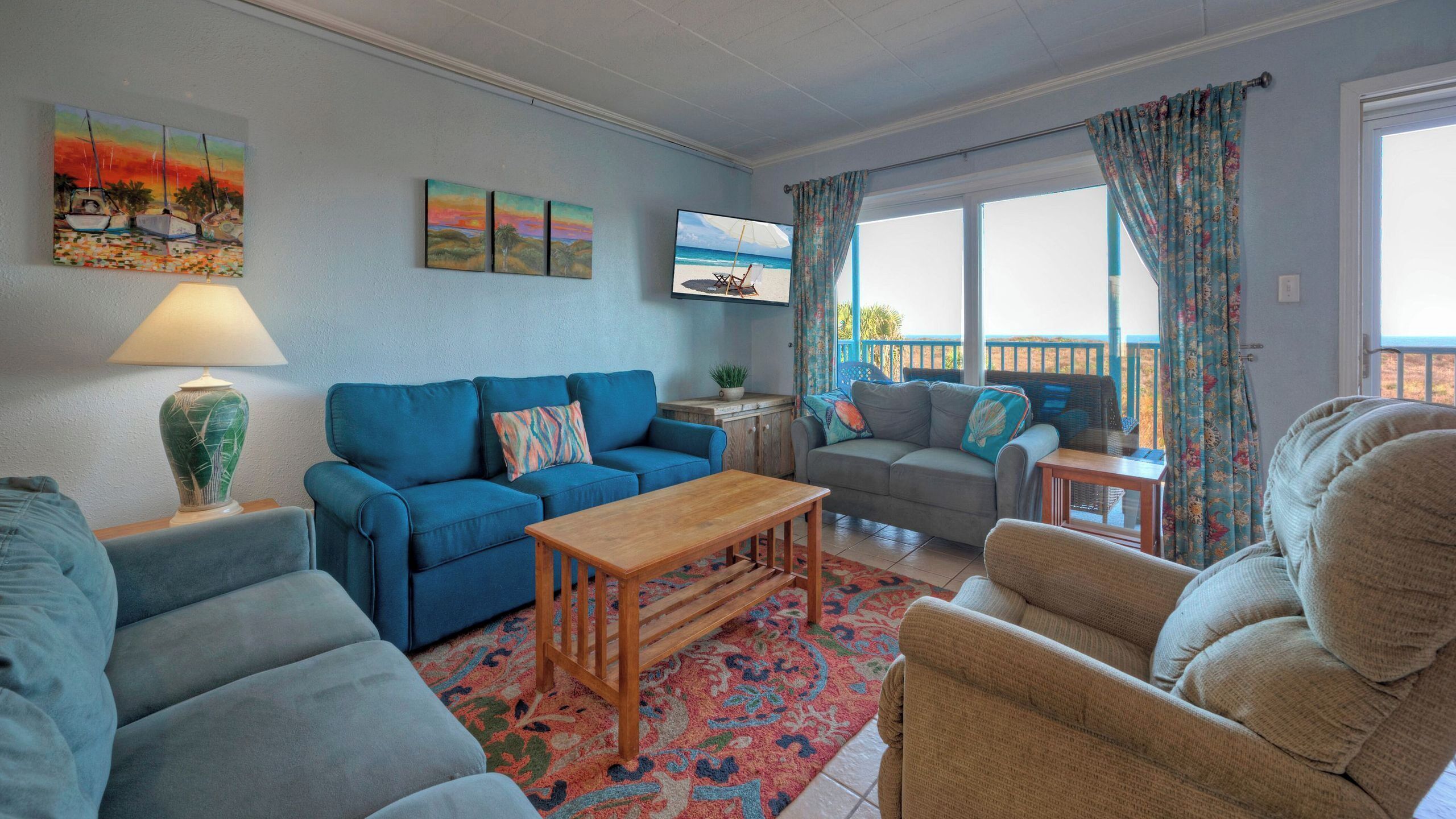 Ein Wohnzimmer mit einem blauen Sofa, einem grauen Sofa, einem Holztisch und einem beigefarbenen Sessel. An der Wand ist ein Fernseher angebracht, auf einem Beistelltisch steht eine Lampe und es gibt ein Fenster mit Vorhängen.