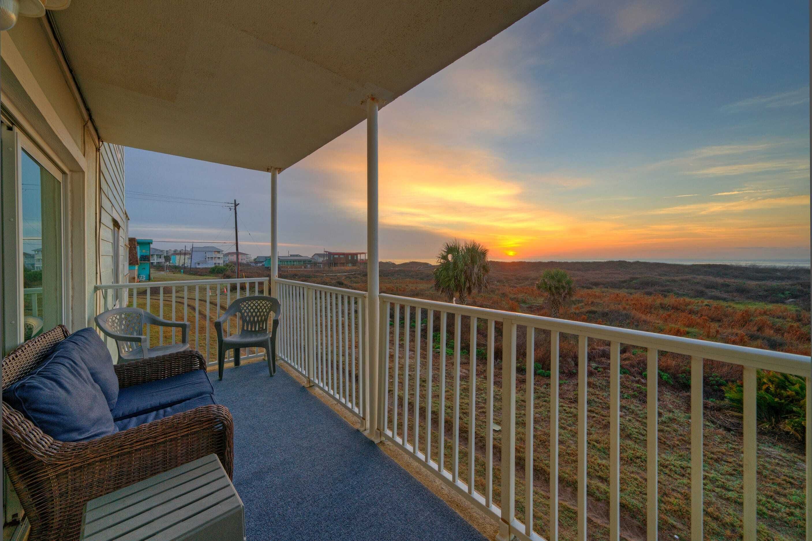 A balcony with a wicker sofa and chairs, overlooking a landscape with a sunset.