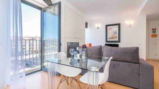Un salon moderne avec une table à manger en verre, deux chaises blanches, un canapé gris et une vue sur la ville à travers de grandes fenêtres.