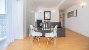 Un salon moderne avec une table à manger en verre, deux chaises blanches, un canapé gris et une grande fenêtre ornée de rideaux légers.