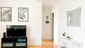 Un salon avec un téléviseur à écran plat posé sur un meuble noir, deux œuvres d’art encadrées représentant des feuilles tropicales, une console blanche avec un miroir et un couloir menant à une sortie.