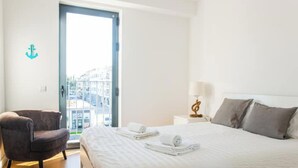 Une chambre d’hôtel avec un lit, une chaise, une table de chevet et une vue sur les immeubles, visible à travers la porte.