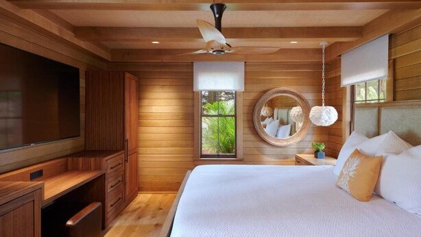 A bedroom with a large bed, wooden walls, a ceiling fan, a TV, and a window with a view of greenery. - Hotel Lanai (Lanai City)
