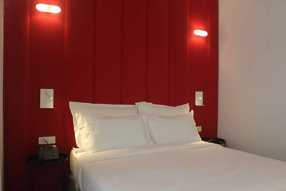 A hotel room with a red accent wall, a bed with white linens, a bedside table with a phone, and a nightstand with a lamp.