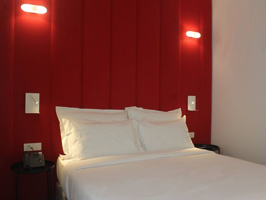 A hotel room with a red accent wall, a bed with white linens, a bedside table with a phone, and a nightstand with a lamp.