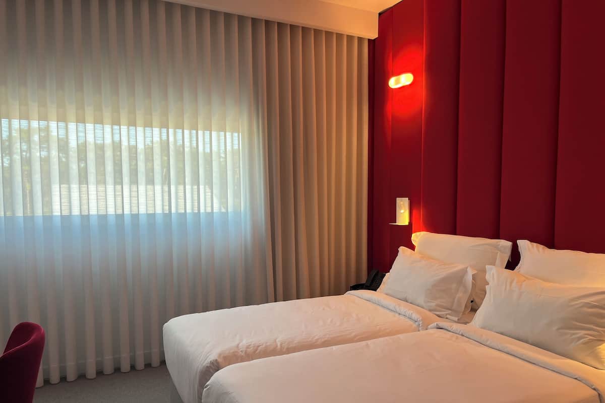 A hotel room with two beds, a red accent wall, a nightstand, and a window with sheer curtains.