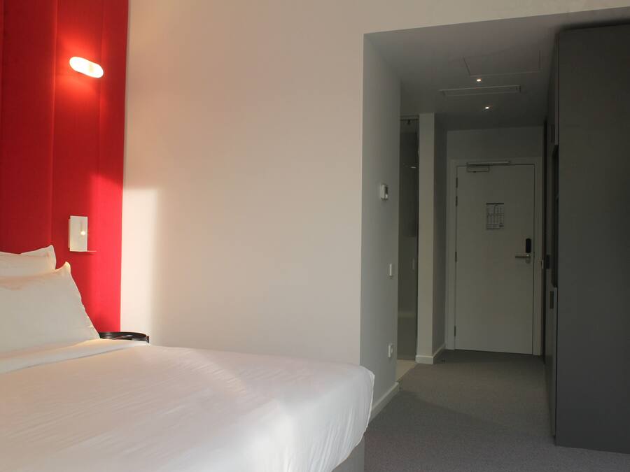 A hotel room with a bed, a red accent wall, and a door leading to another room.