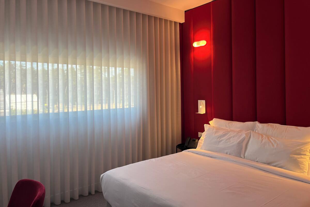 A hotel room with a large bed, white bedding, a red accent wall, a bedside table with a phone, and sheer curtains allowing natural light.