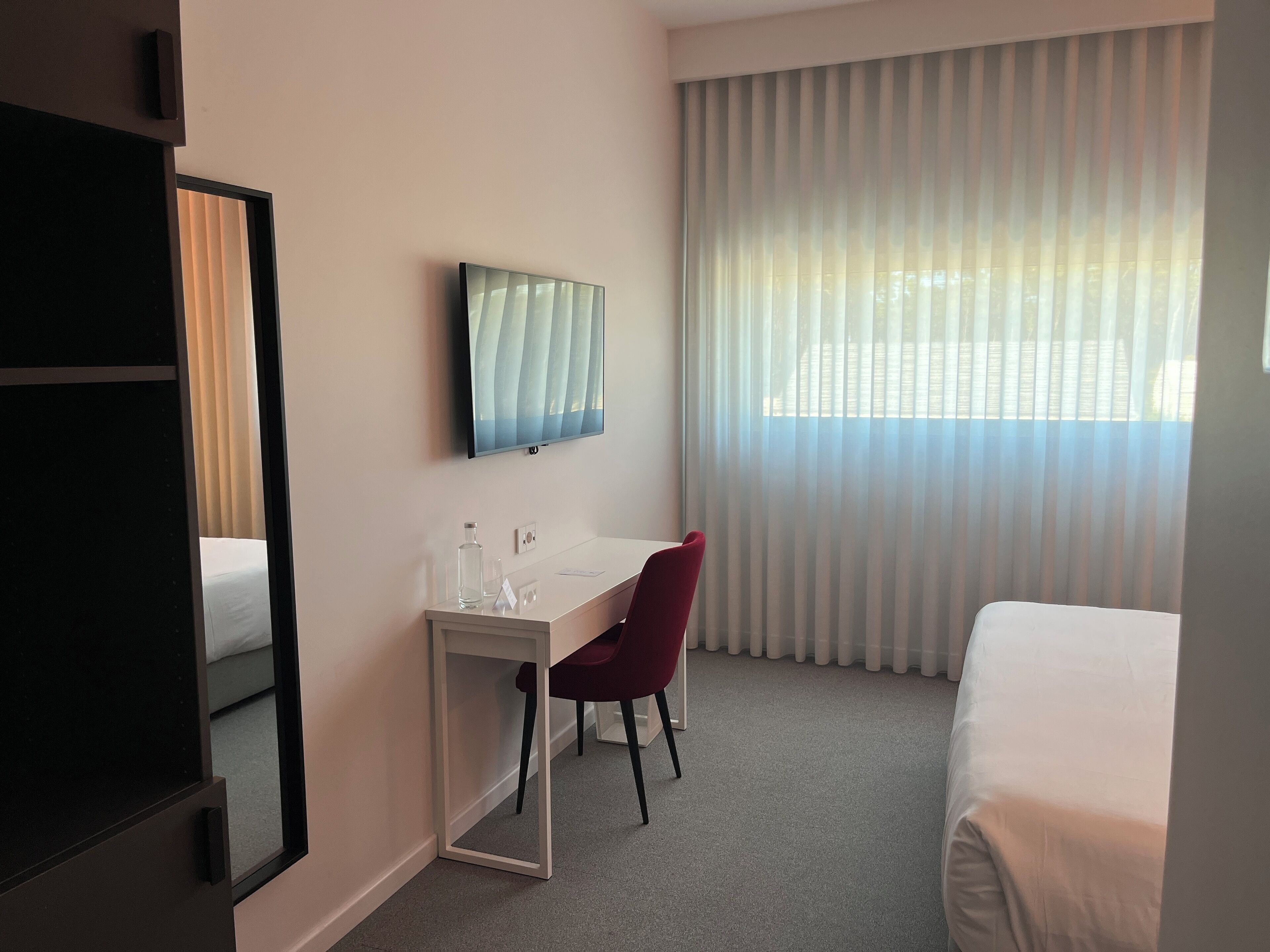 Une chambre d’hôtel équipée d’un lit, d’un bureau, d’une chaise, d’une télévision et d’une fenêtre avec des stores.